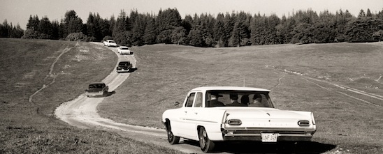 A caravan of 60's era cars containing UC Regents drives up a dirt road.